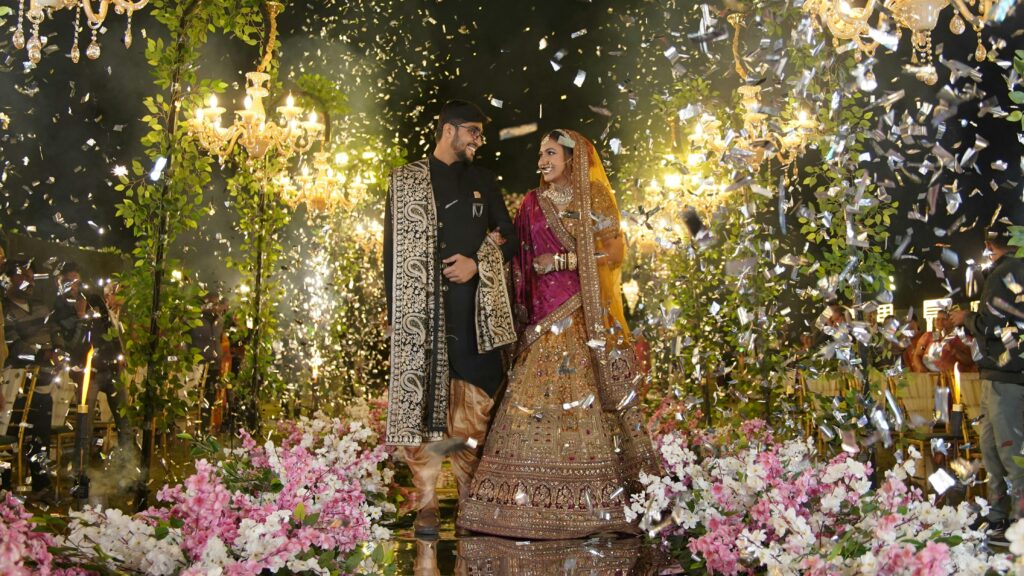 A joyful Indian bride and groom at a vibrant outdoor wedding with decorative lighting and flowers.